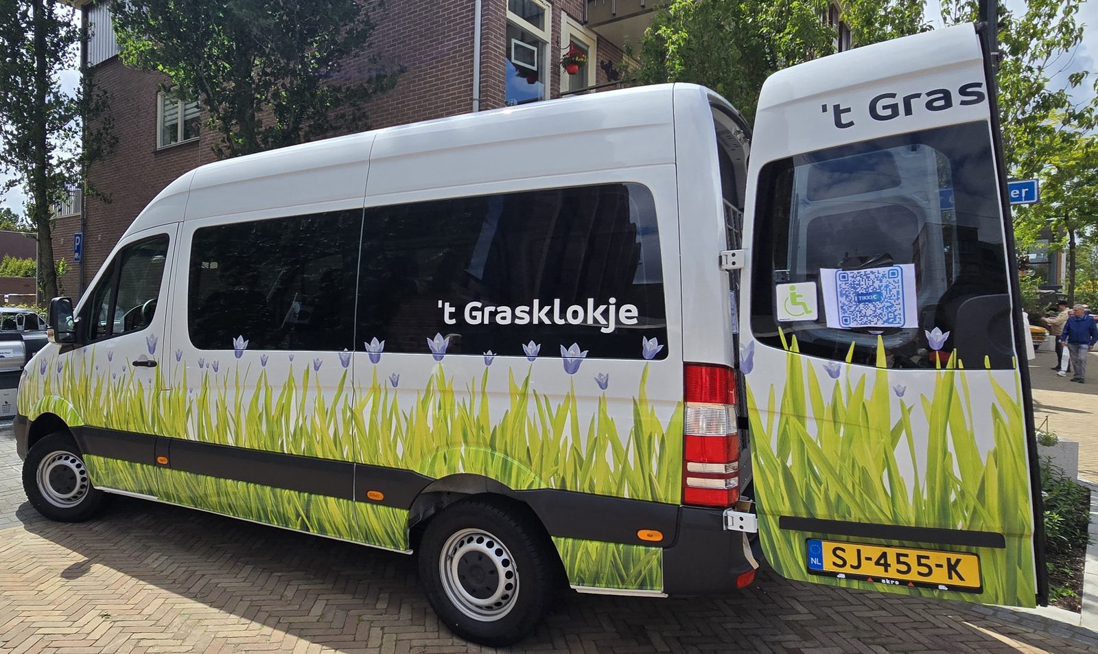 De rolstoelbus van 't Grasklokje van opzij en achter, met logo, het woord 't Grasklokje en kenteken